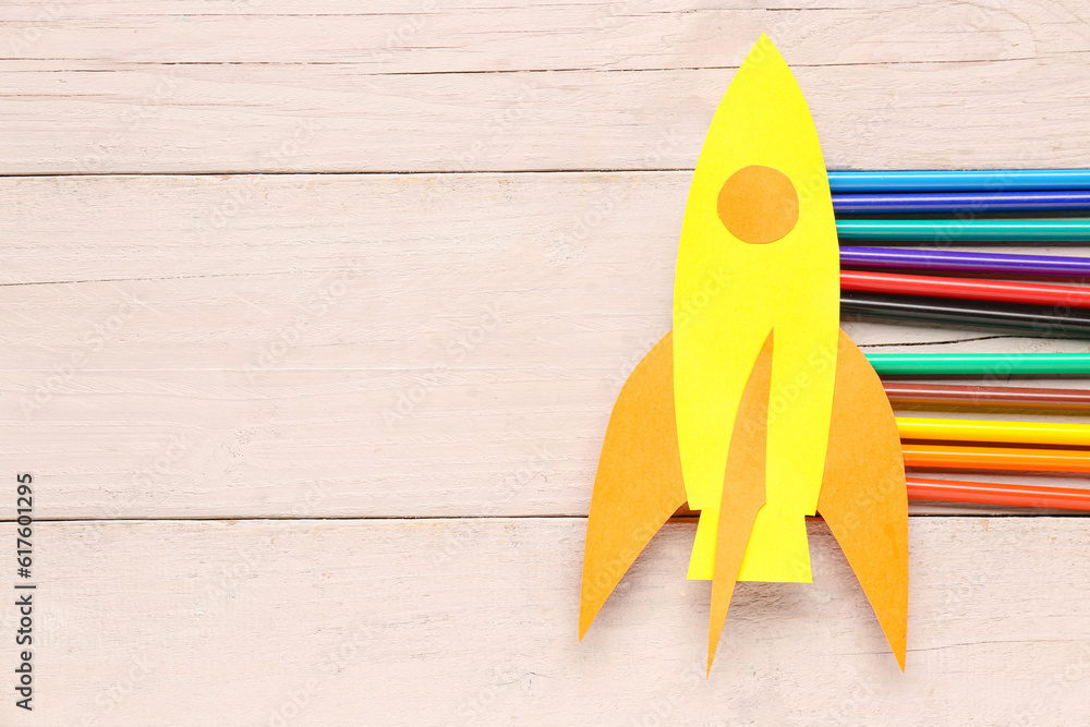 Yellow paper rocket with color pencils on beige wooden background