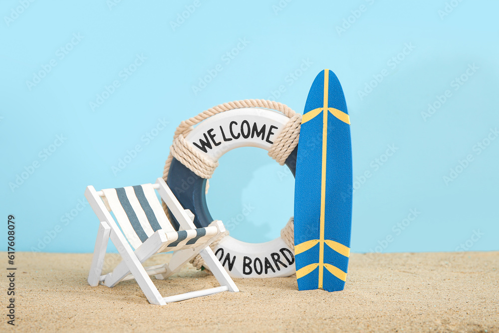 Creative composition with miniature surfboard, deckchair and life buoy on sand against blue backgrou