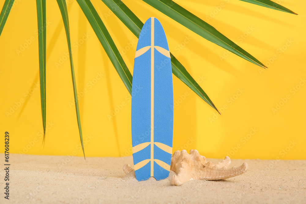 Mini surfboard with starfish and palm leaf on sand against yellow background