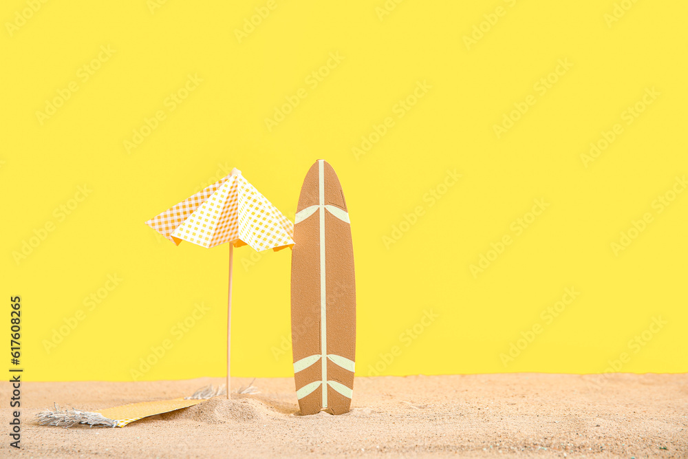 Creative composition with mini surfboard, umbrella and blanket on sand against yellow background