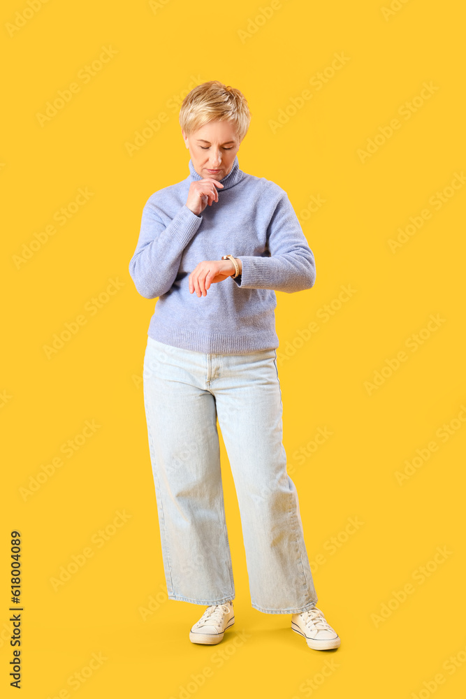 Thoughtful mature woman looking at wristwatch on yellow background