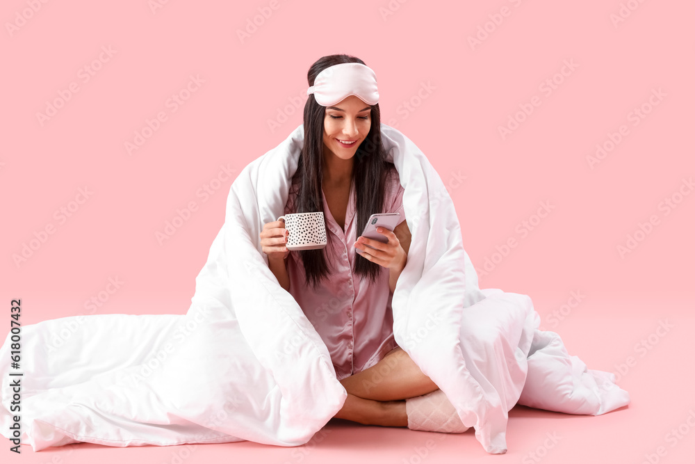 Young happy woman with cup of coffee and blanket sitting and using mobile phone on pink background