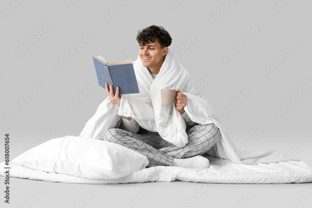 Handsome happy man with cup of coffee, soft blanket and pillow reading book on grey background