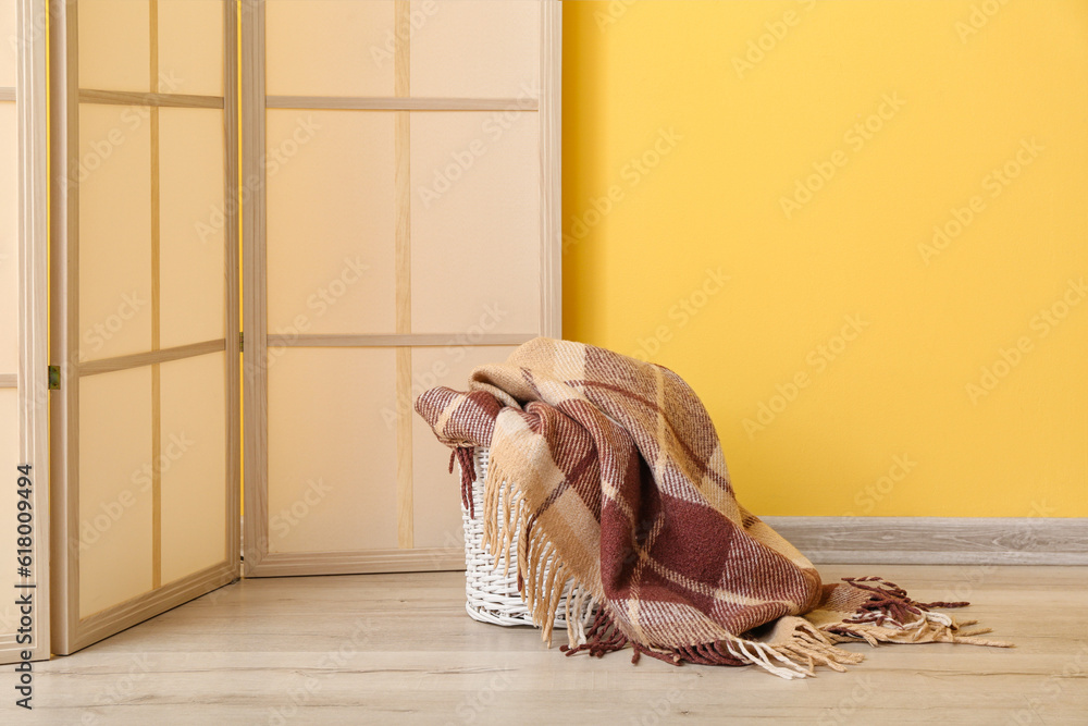 Basket with new soft blanket and folding screen near yellow wall in room