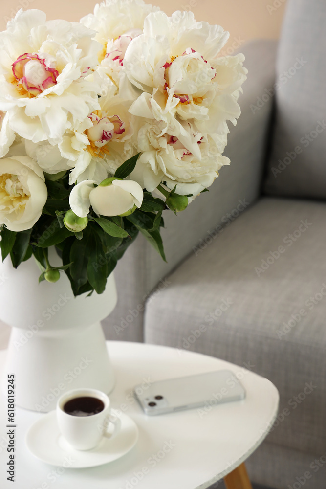 Vase of white peonies with cup of coffee and mobile phone on table near couch