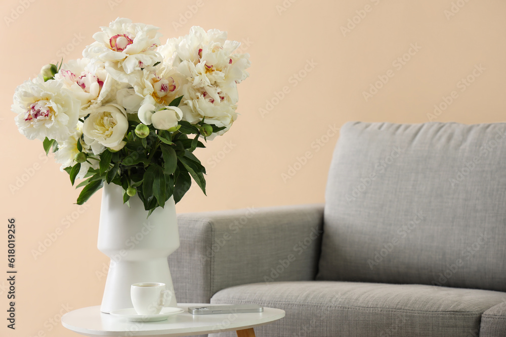 Vase of white peonies with cup of coffee and mobile phone on table near couch