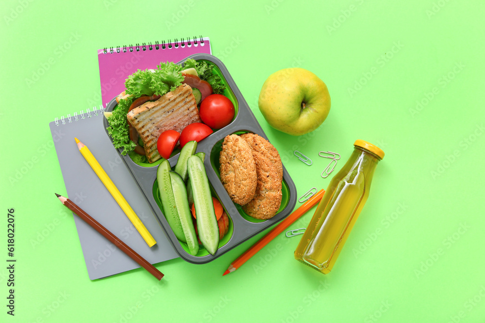 Stationery, drink and lunch box with tasty food on green background