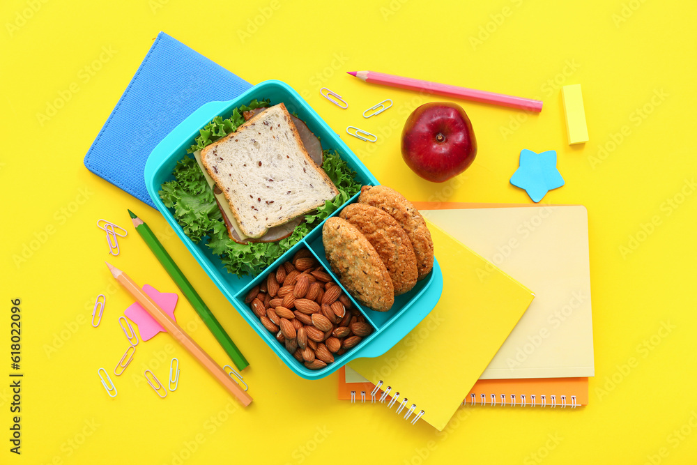 Stationery and lunch box with tasty food on yellow background