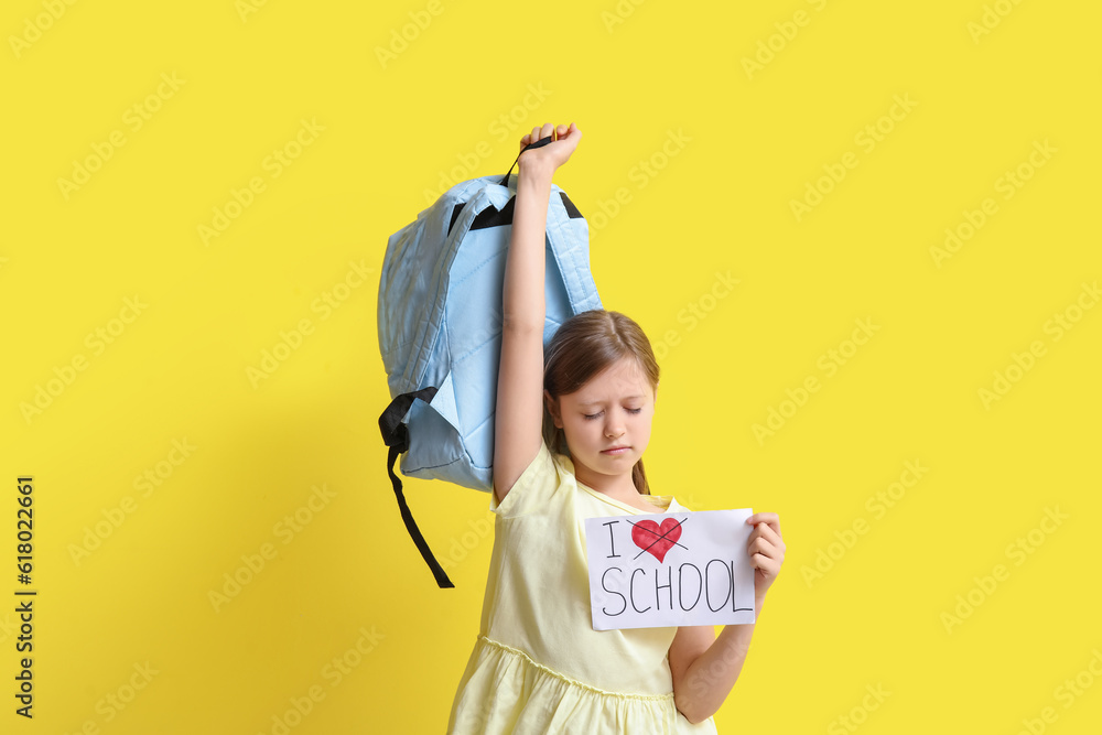 Little girl holding paper with text I DO NOT LOVE SCHOOL and backpack on yellow background