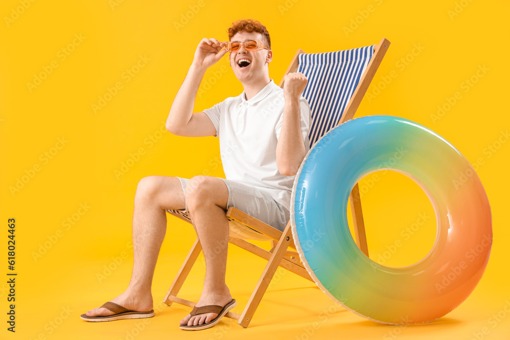 Young man with inflatable ring resting in deck chair on yellow background