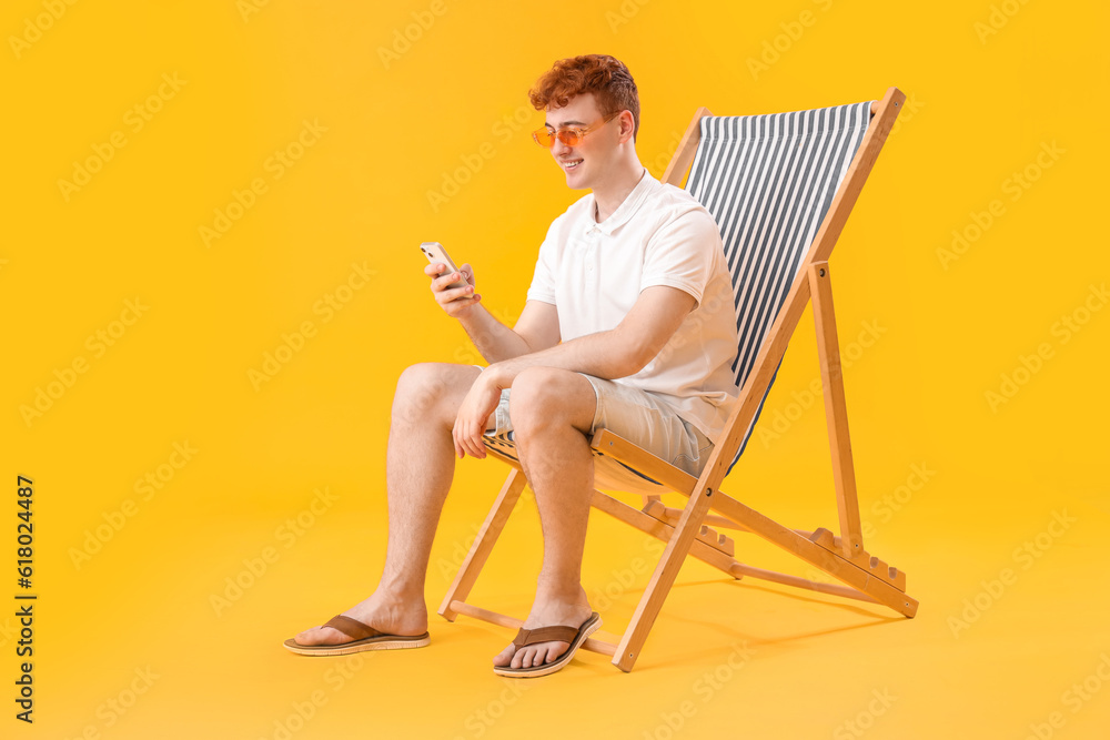 Young man using mobile phone in deck chair on yellow background