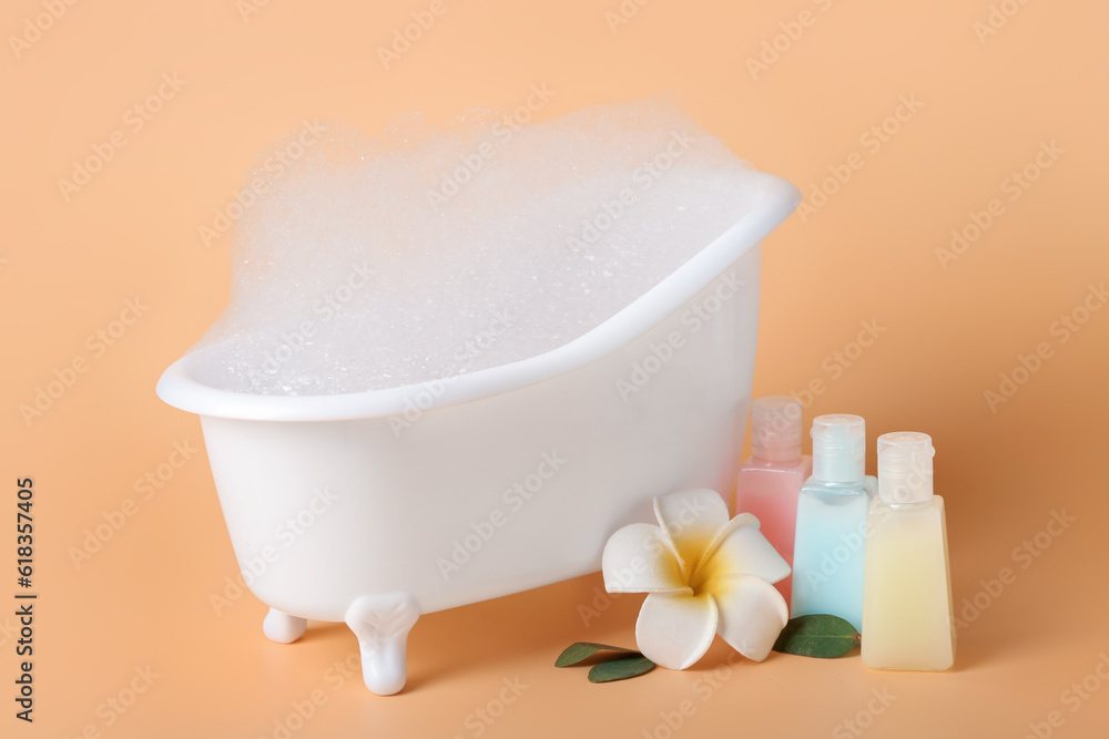 Small bathtub with foam and bottles of cosmetic products on color background