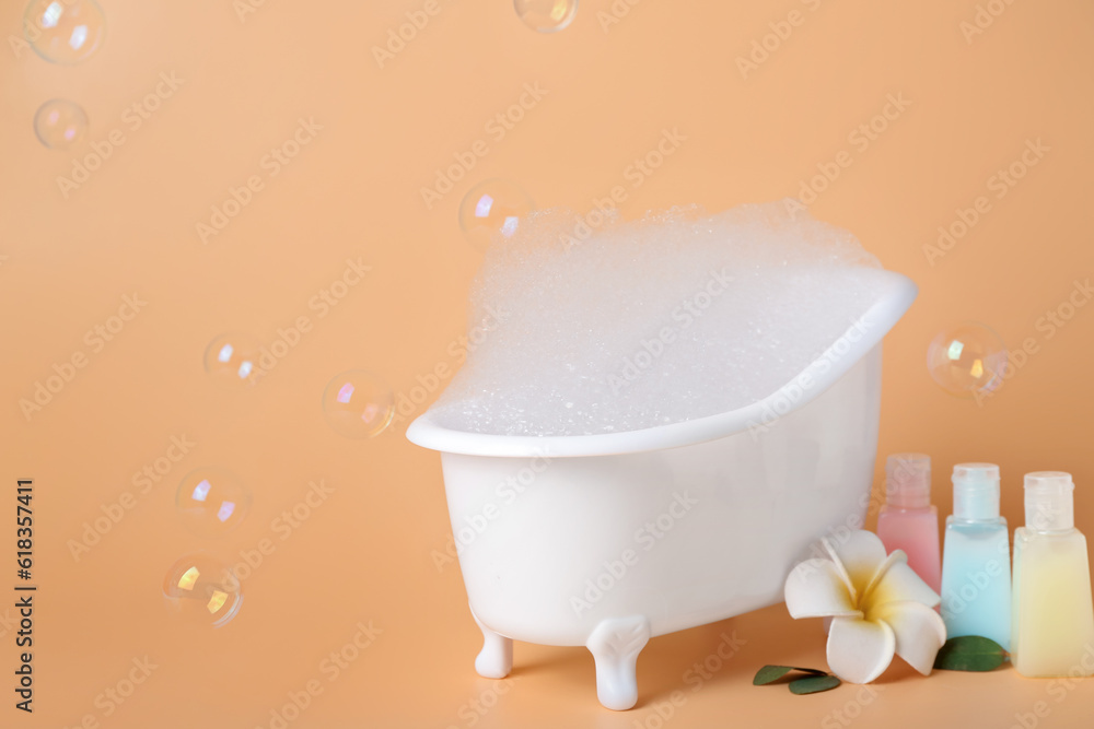 Small bathtub with foam, soap bubbles and bottles of cosmetic products on color background