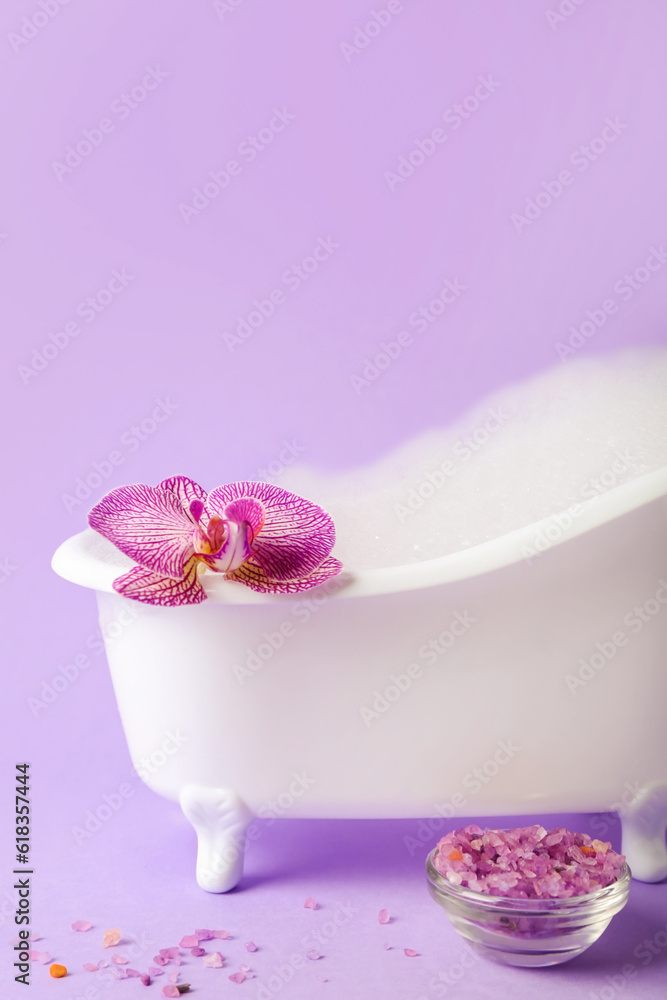 Small bathtub with foam, sea salt and orchid flower on lilac background