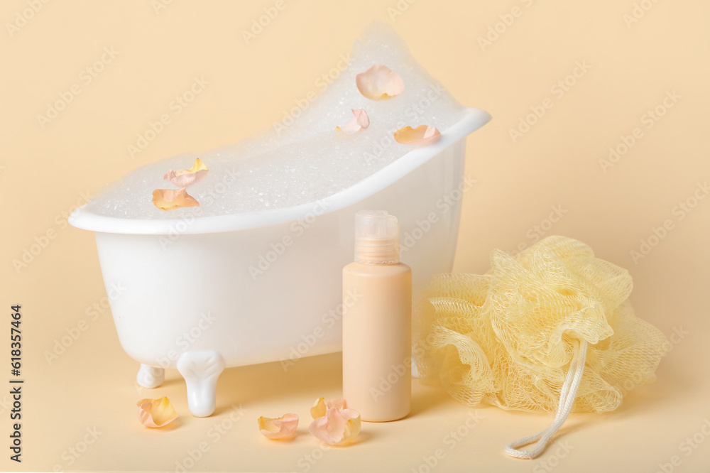 Small bathtub with foam, flower petals, sponge and bottle of cosmetic product on color background