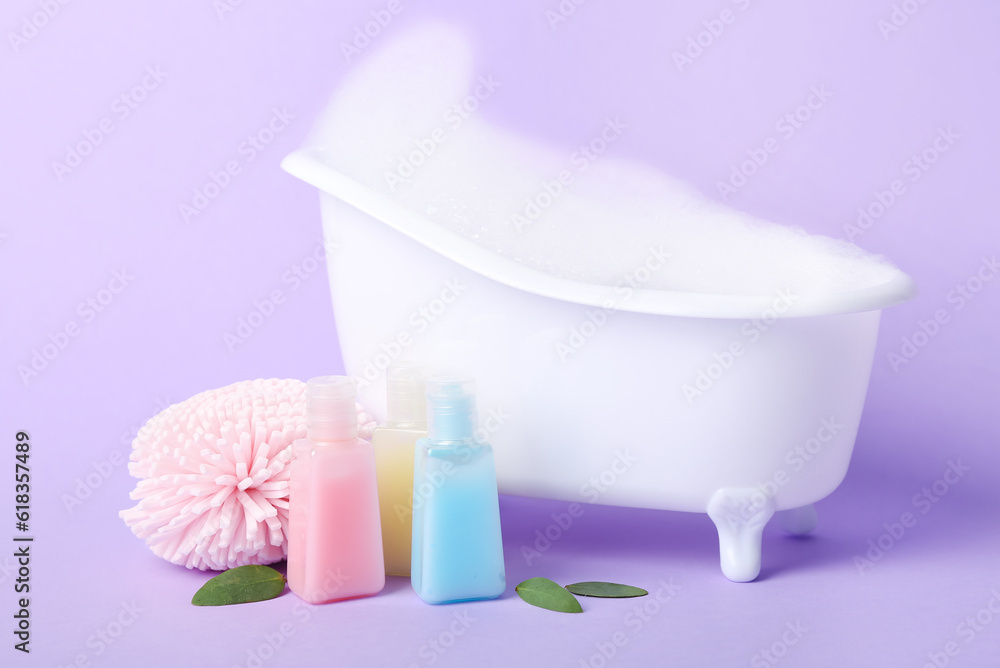 Small bathtub with foam, sponge and bottles of cosmetic products on lilac background