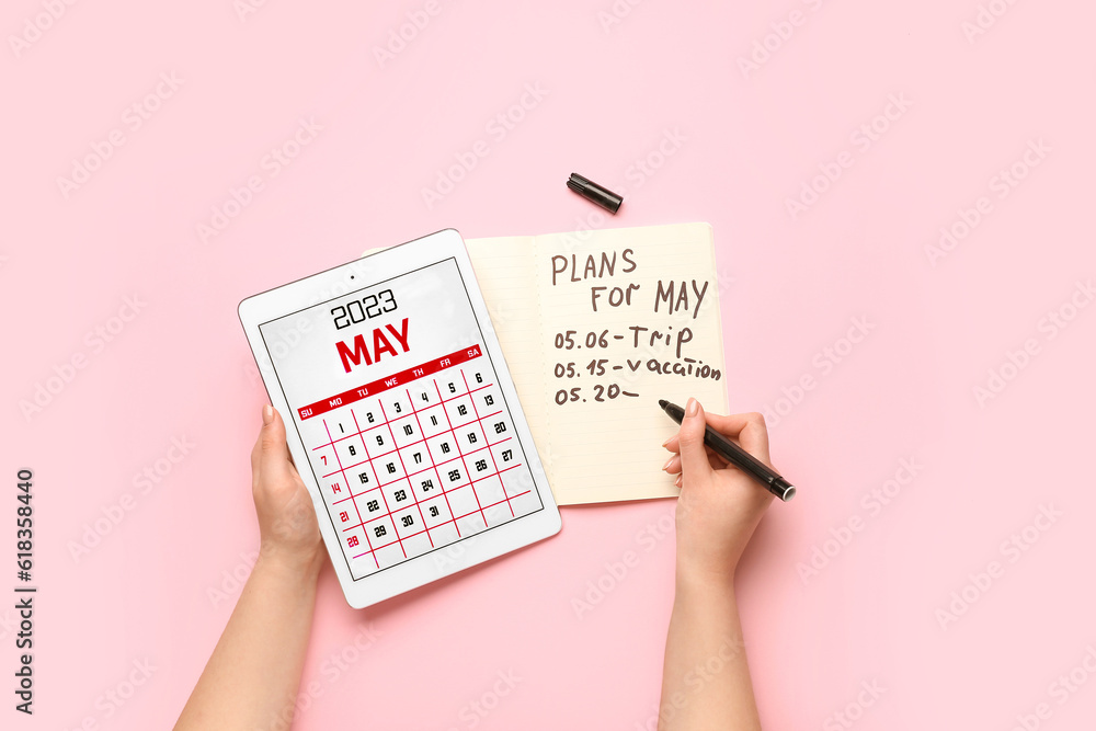 Woman holding tablet computer with calendar and writing plans for May on pink background