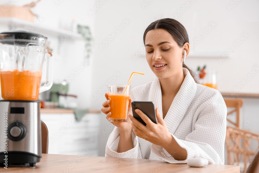 Young woman with glass of vegetable juice using mobile phone in kitchen