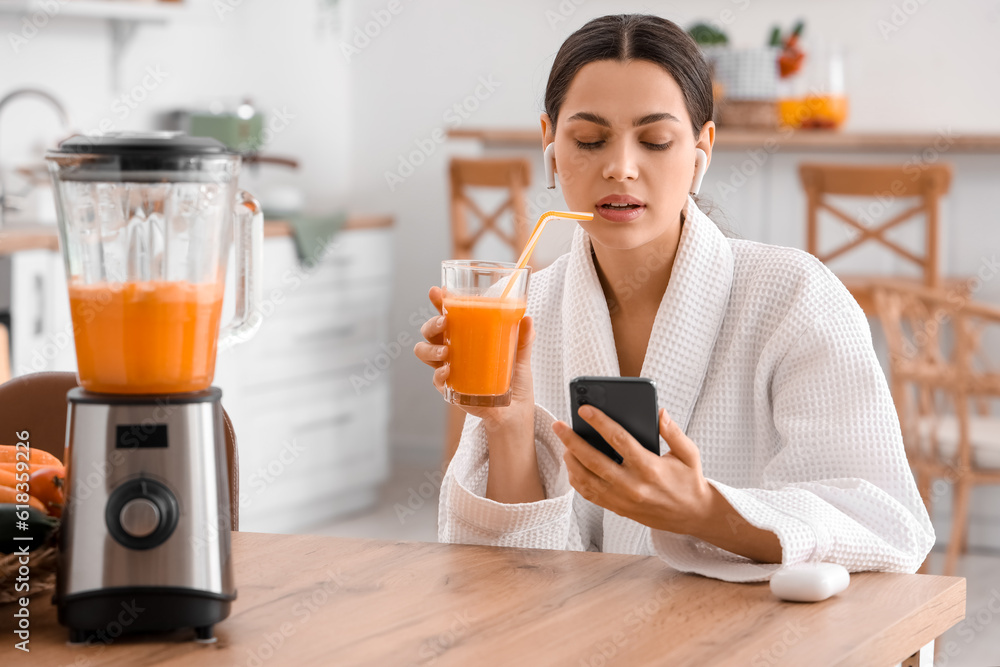 Young woman with glass of vegetable juice using mobile phone in kitchen