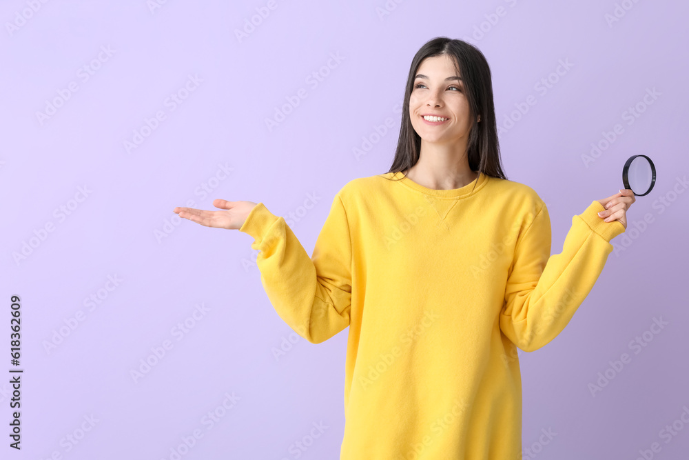 Beautiful happy young woman with magnifier pointing at something on lilac background