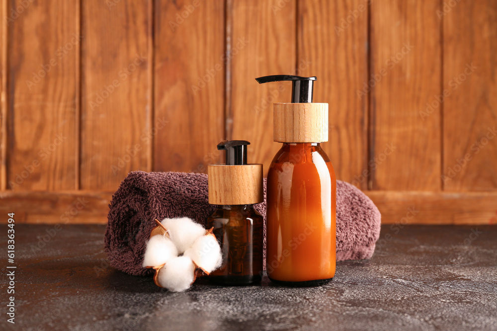 Bottles of cosmetic products, clean towel and cotton flower on dark table against wooden background