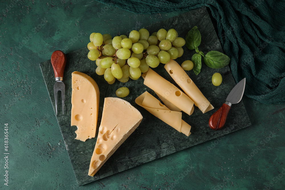 Board with tasty Swiss cheese and grapes on green background