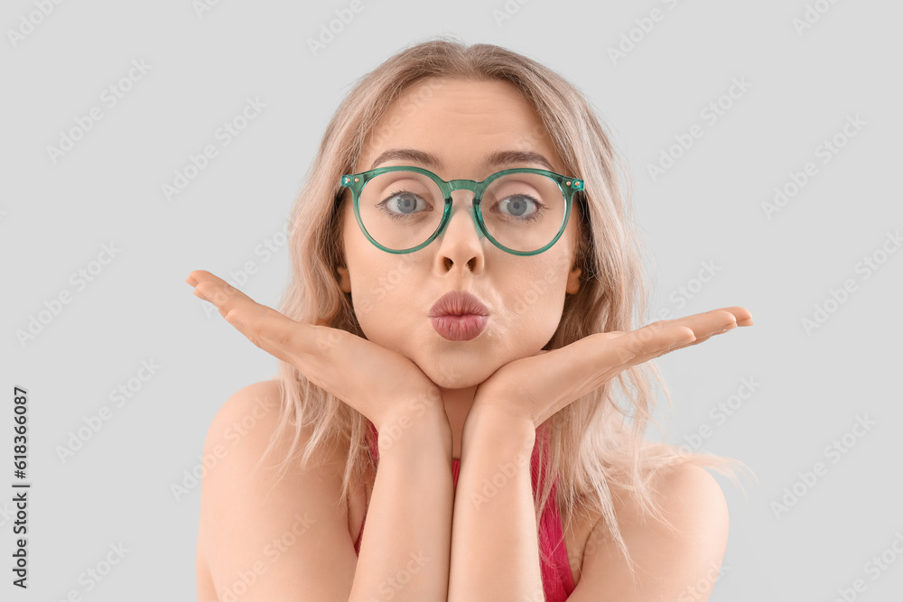 Young woman in stylish eyeglasses blowing kiss on light background, closeup