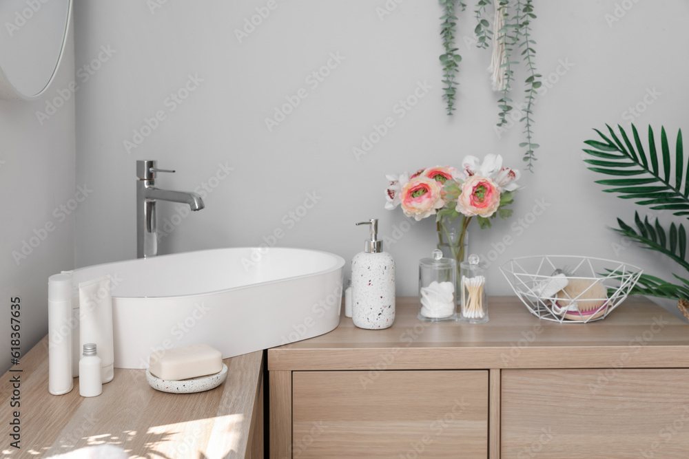 Sink bowl, bath accessories and ranunculus flowers on wooden cabinet in light bathroom