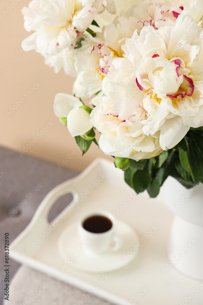 Vase of white peonies with cup of coffee on tray near beige wall