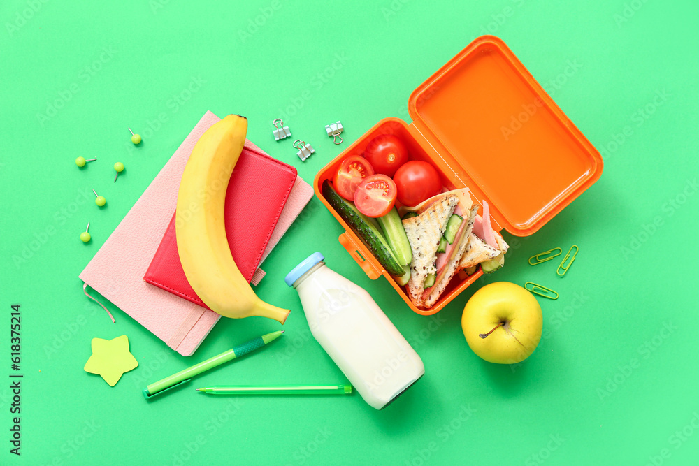 Stationery, drink and lunch box with tasty food on green background