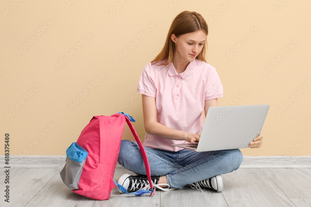 Female student with backpack using laptop near beige wall