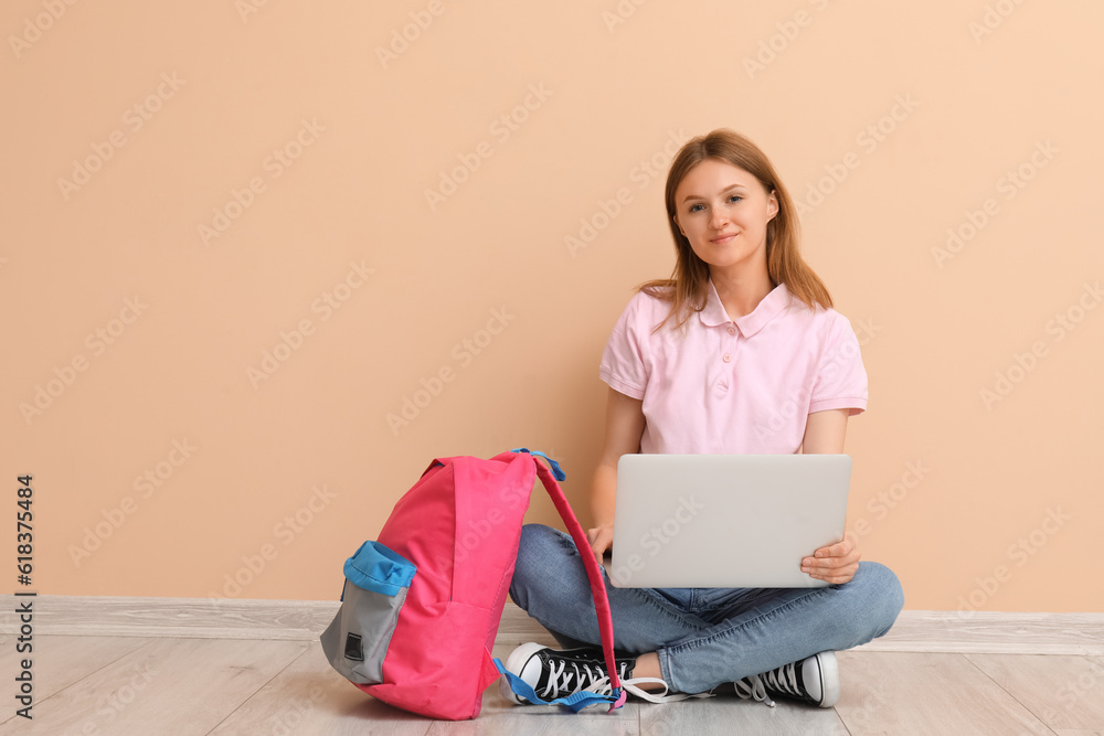 Female student with backpack using laptop near beige wall