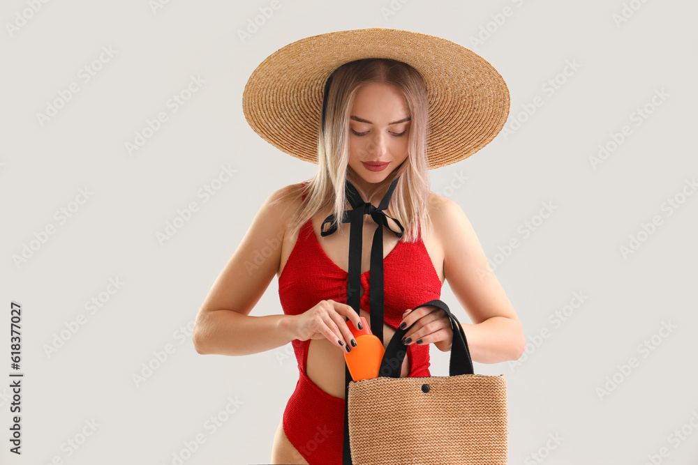 Young woman putting sunscreen cream in bag on light background