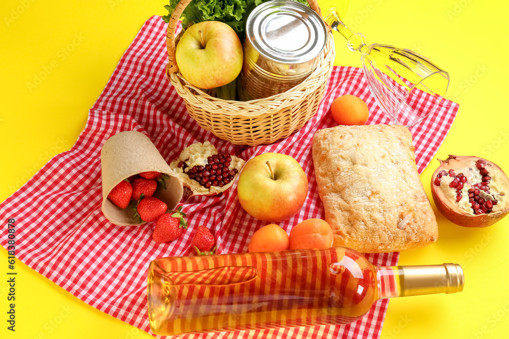 Wicker basket with tasty food for picnic, wine and glass on yellow background, closeup