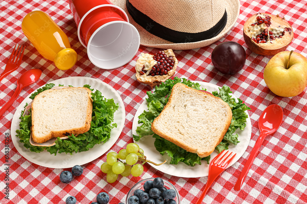 Composition with hat, plastic tableware, juice and sandwiches for picnic on fabric background, close