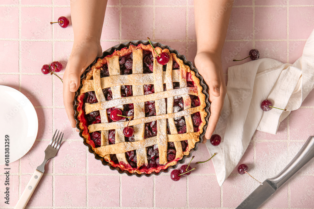 Female hands holding baking dish with tasty cherry pie on pink tile background