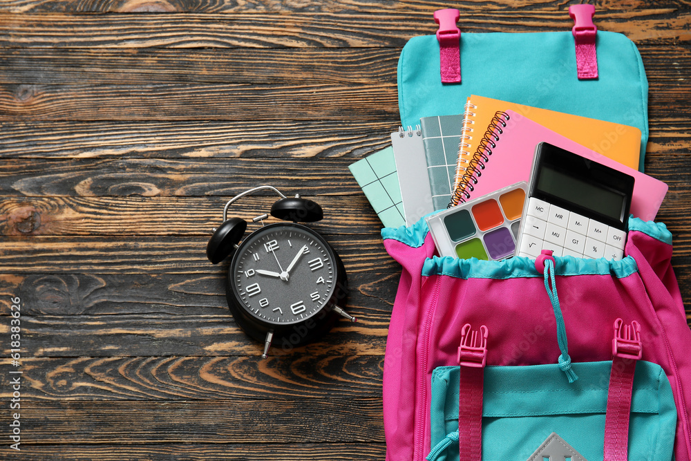 Colorful school backpack with alarm clock, notebooks and calculator on brown wooden background