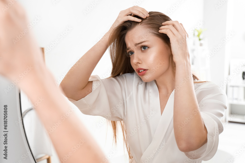 Worried young woman with hair loss problem looking in mirror at home