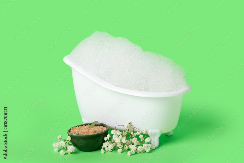 Small bathtub with foam, sea salt and gypsophila flowers on green background