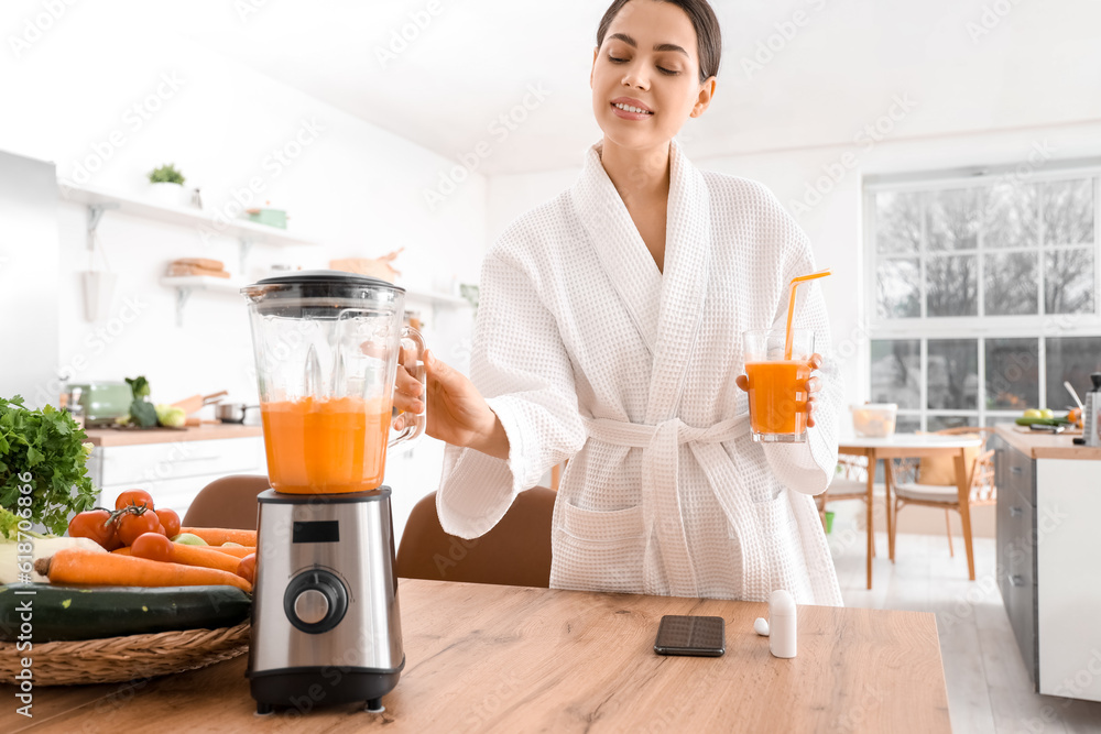 Young woman with glass of vegetable juice and blender in kitchen