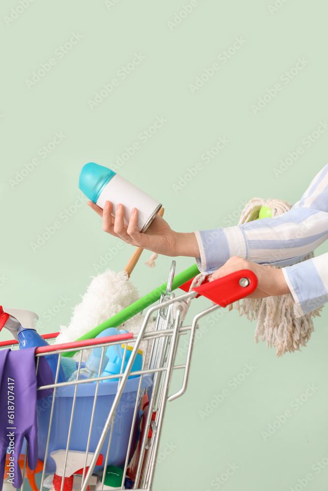 Woman with shopping cart full of cleaning supplies on pale green background