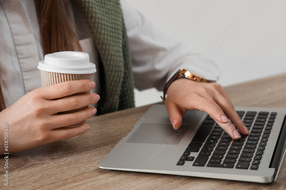 Woman using modern laptop and holding takeaway cup of coffee at table