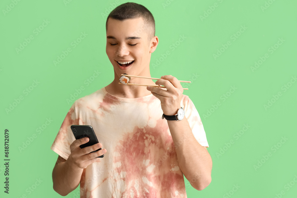 Young man with tasty sushi roll and mobile phone on green background