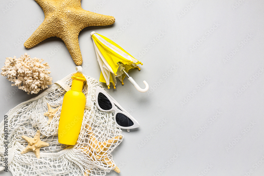 String bag with sunglasses, mini umbrella, coral and bottle of sunscreen cream on grey background