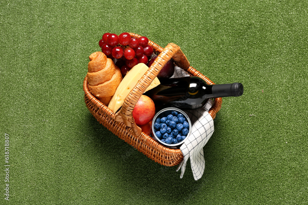 Wicker basket with tasty food for picnic and wine on green background