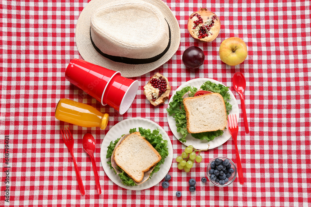 Composition with hat, plastic tableware, juice and sandwiches for picnic on fabric background