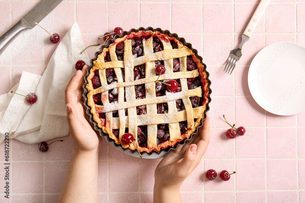 Female hands holding baking dish with tasty cherry pie on pink tile background