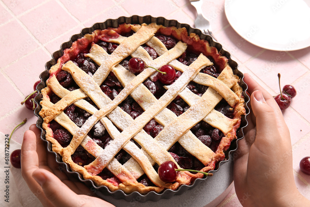 Female hands holding baking dish with tasty cherry pie on pink tile background