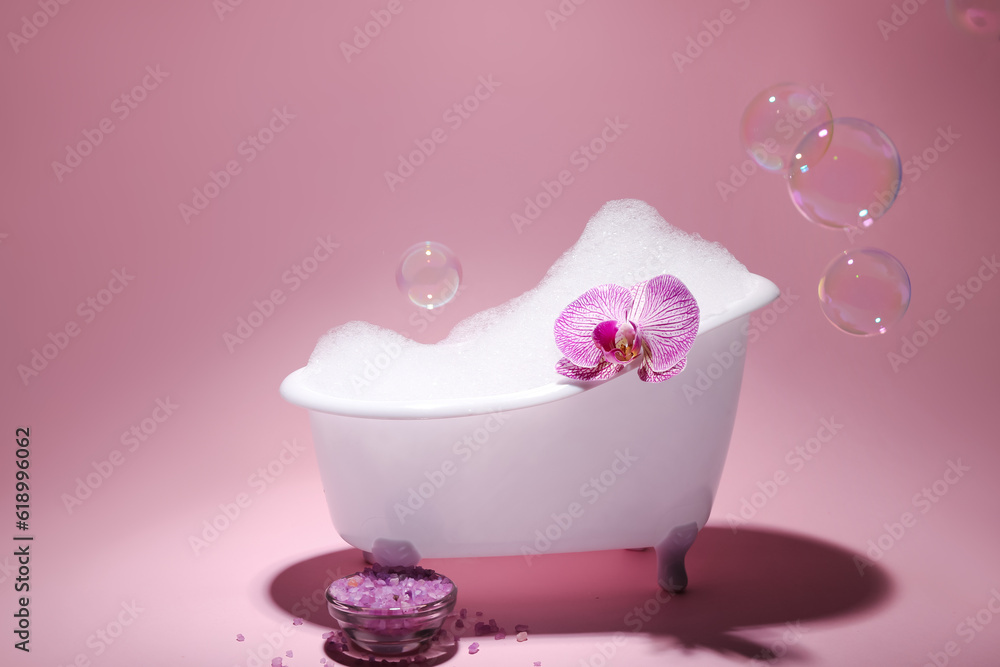 Small bathtub with foam, soap bubbles, sea salt and orchid flower on pink background