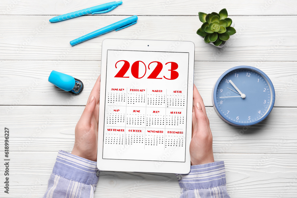 Woman holding tablet computer with calendar near alarm clock, stapler and houseplant on white wooden